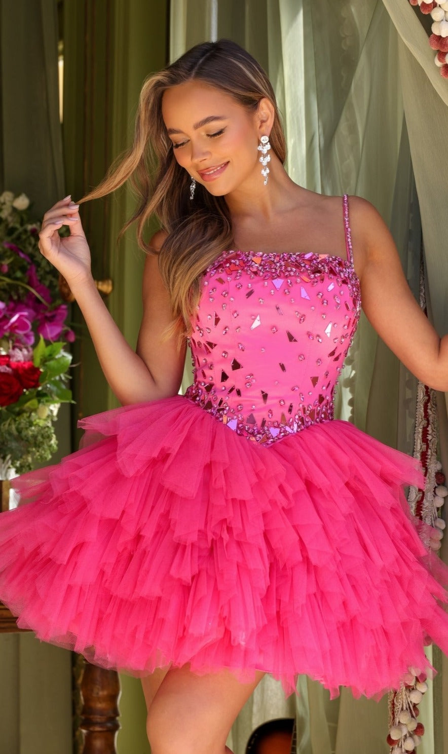 A woman smiles in the Ava Presley 29180 short tiered homecoming dress, featuring bright pink beading and a layered tulle skirt. She wears earrings and is surrounded by green curtains and flowers.